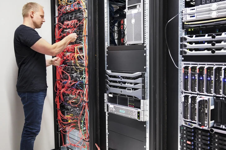 Un technicien informatique travaille dans une salle serveur, utilisant un ordinateur portable pour administrer l'infrastructure