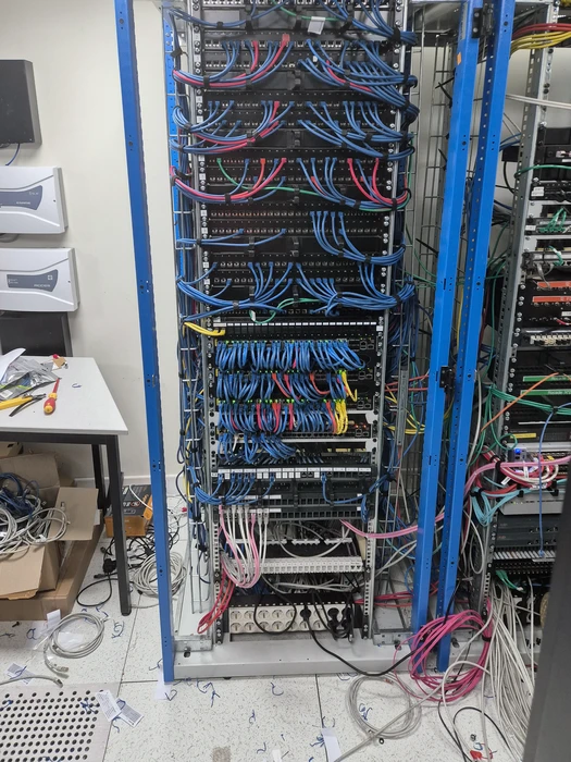 Salle informatique en cours d'installation, baie de brassage 42U à montants bleus, dense câblage structuré : câbles RJ45 Cat6 bleus, roses, jaunes, blancs, patch panels 24 ports, switches managés, PDU verticale, chemins de câbles métalliques