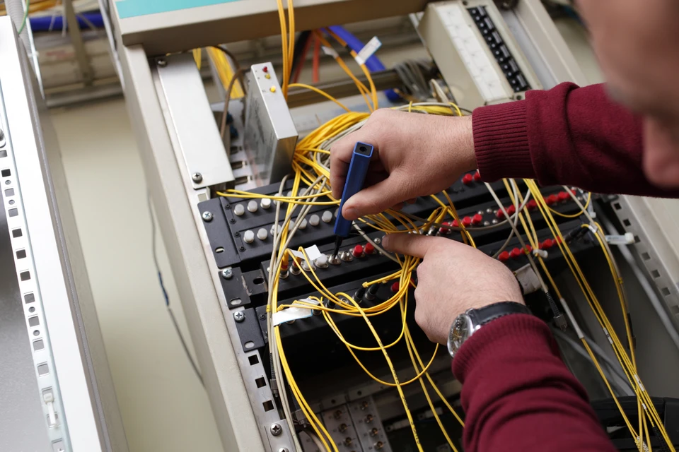 Intérieur d'un centre de données, technicien en pull bordeaux manipulant des câbles de fibre optique jaunes dans une baie de brassage réseau