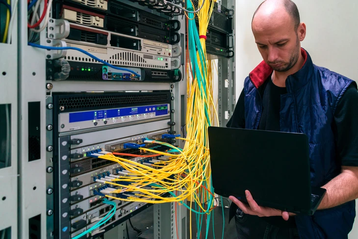 Un technicien informatique travaille dans une salle de serveurs, agenouillé devant des baies de serveurs ouvertes