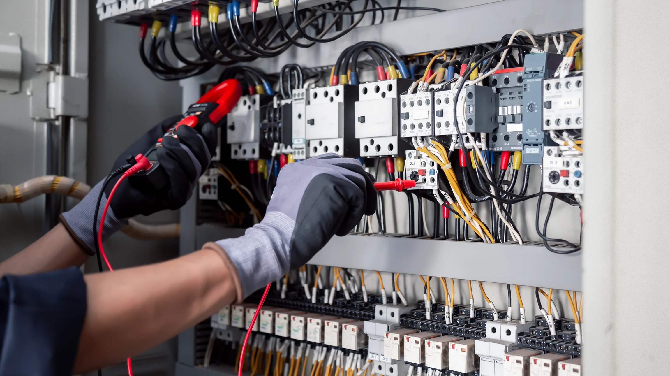 Technicien en uniforme bleu effectuant une mesure électrique dans un tableau de distribution domestique avec un multimètre numérique orange