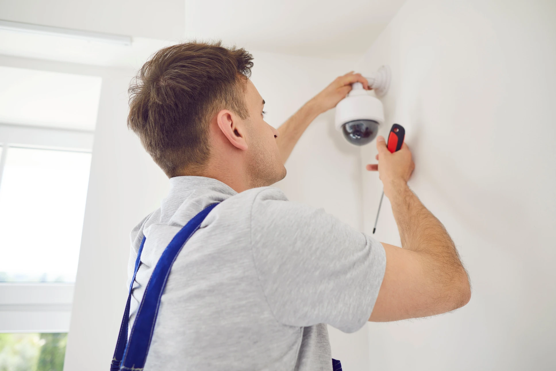 Un technicien en uniforme rouge installe une caméra de surveillance au plafond d'un intérieur aux murs blancs